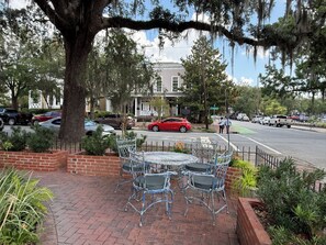 A paved outdoor seating area with metal chairs and tables, surrounded by brick planters and a large tree. - Savannah Manor- Grand Historic Victorian Mansion (Savannah)