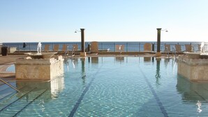 Piscina cristalina con vista al océano, rodeada de sillas de descanso y una barandilla.