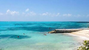Une plage à l’eau turquoise et limpide, avec un rivage sablonneux et quelques personnes qui se baignent.