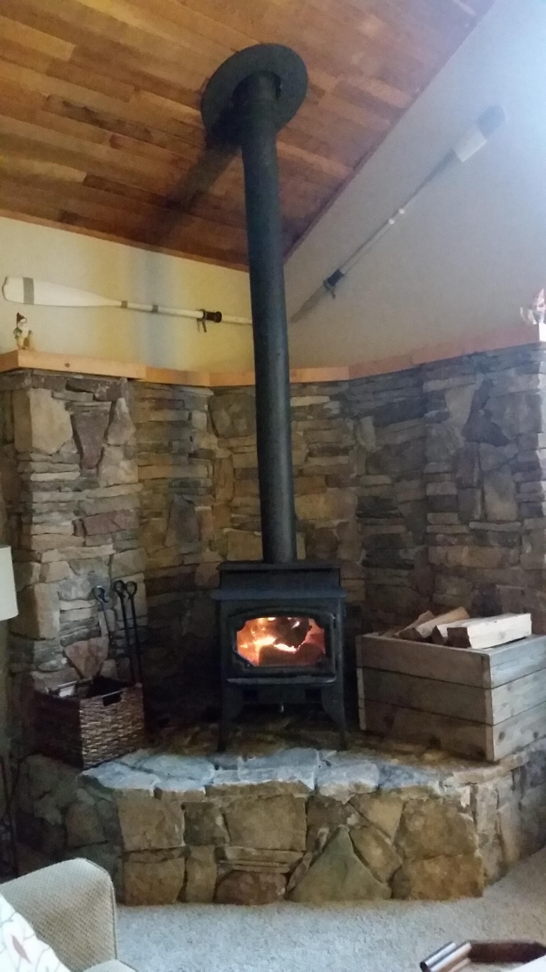 A stone fireplace with a black chimney, a fire burning inside, and a stone wall surrounding it.