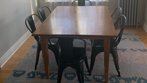 Un coin repas avec une table en bois et six chaises, deux fenêtres avec des rideaux légers, un radiateur et un tapis à motifs.