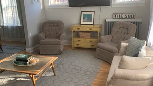 Un salon avec un ventilateur de plafond, deux fauteuils, un canapé, une table basse avec des livres, un téléviseur et un tableau encadré posé sur une étagère.