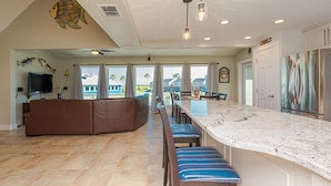A spacious kitchen with a large island, a dining area with bar stools, a brown sofa, and a TV mounted on the wall.