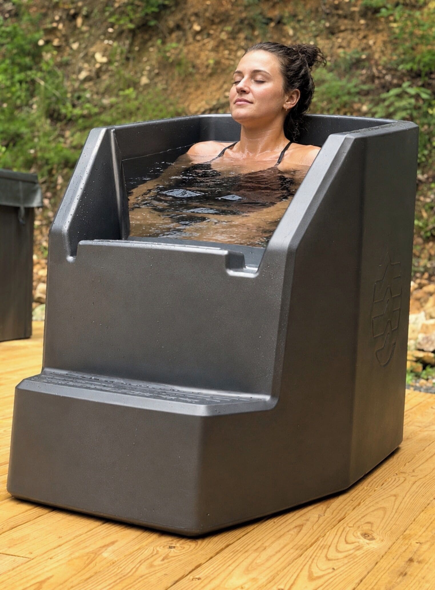 Una persona en una bañera exterior con hidromasaje y una terraza de madera al fondo.