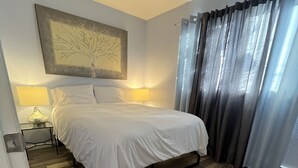 Un dormitorio con piso de madera, una cama con ropa de cama blanca, una mesita de noche con lámpara y un cuadro en la pared.