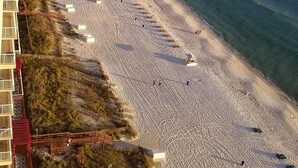 En strand med rækker af liggestole og parasoller, en klar himmel og en bygning med altaner med udsigt over stranden.