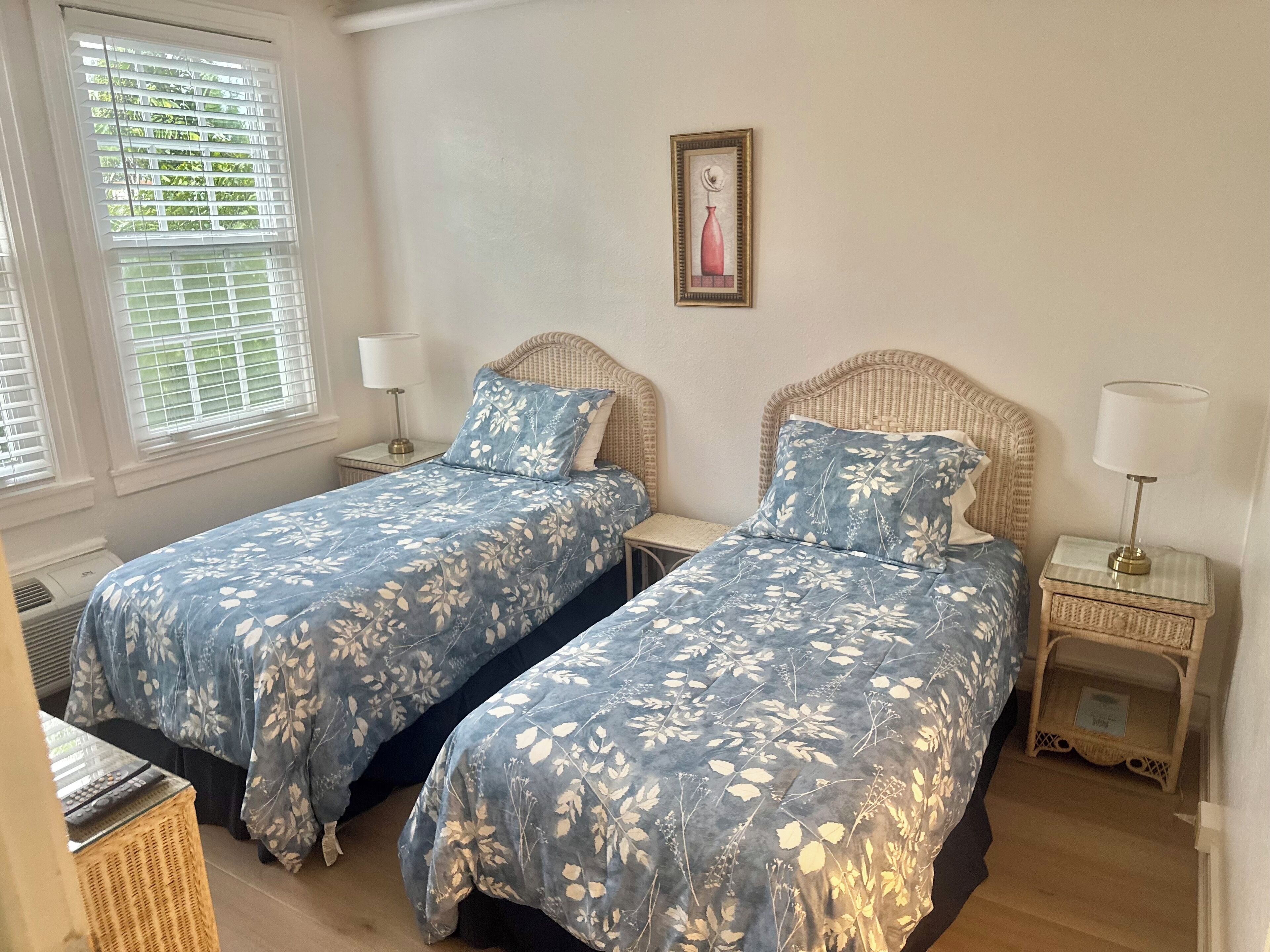 A room with two single beds, each with a matching headboard and bedside table. There is a framed picture on the wall, and the room has a window with blinds.