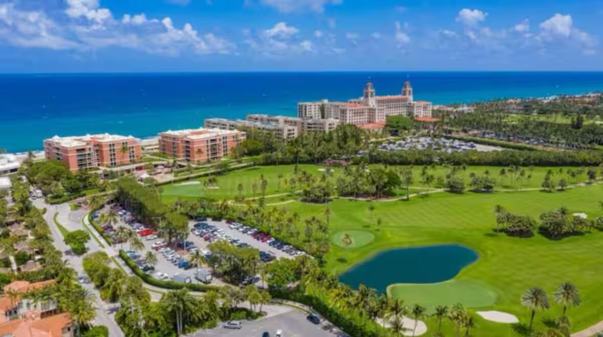 Vista aerea di un resort con diversi edifici, un campo da golf e una vasta distesa d'acqua sullo sfondo.
