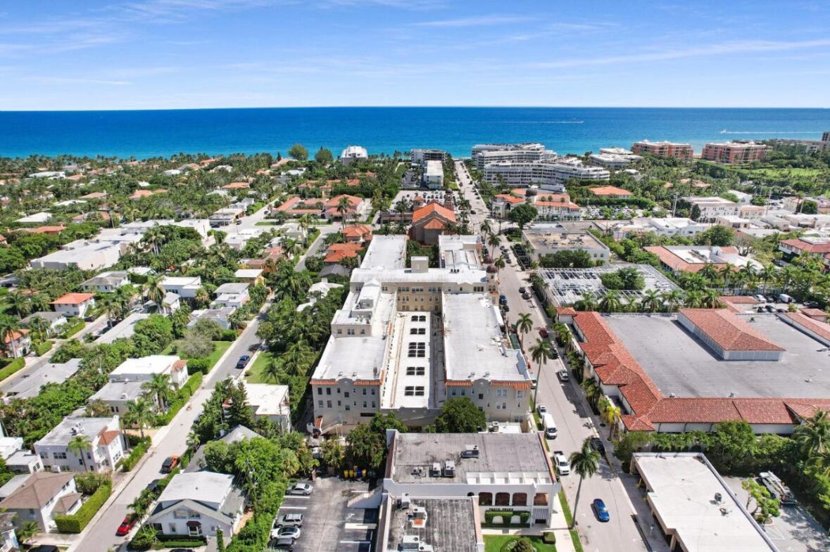 An aerial view of a coastal town with a prominent white building with a red roof in the center, surrounded by residential houses and greenery.