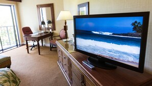 Habitación de hotel con un televisor de pantalla plana grande que muestra una playa, un escritorio de madera con lámpara, una silla y un sofá pequeño.