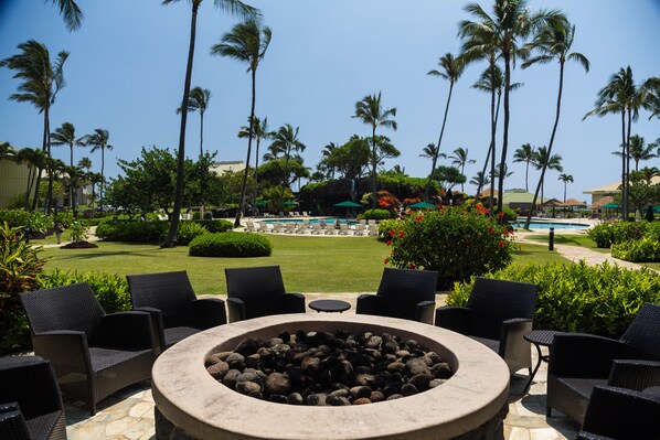 A fire pit surrounded by wicker chairs, overlooking a pool area with lounge chairs and palm trees.