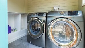A laundry room with two front-loading washing machines, a shelf with laundry supplies, and a power outlet on the wall.