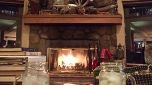 A fireplace with a stone surround, a mantel decorated with antlers and pinecones, and a fire burning. There are two mugs on the mantel, one with ice and a remote control, and a book titled "The Pepper Picture Show" is visible. - Indian Heaven Treehouse-Nightly Shooting Stars, Miles of Balconies & Rock Views  (Idyllwild)