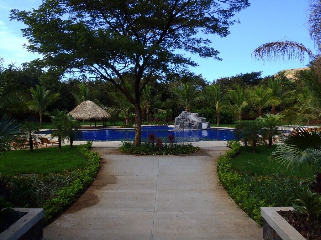 A pathway leading to a pool with a thatched-roof structure and a statue in the center.