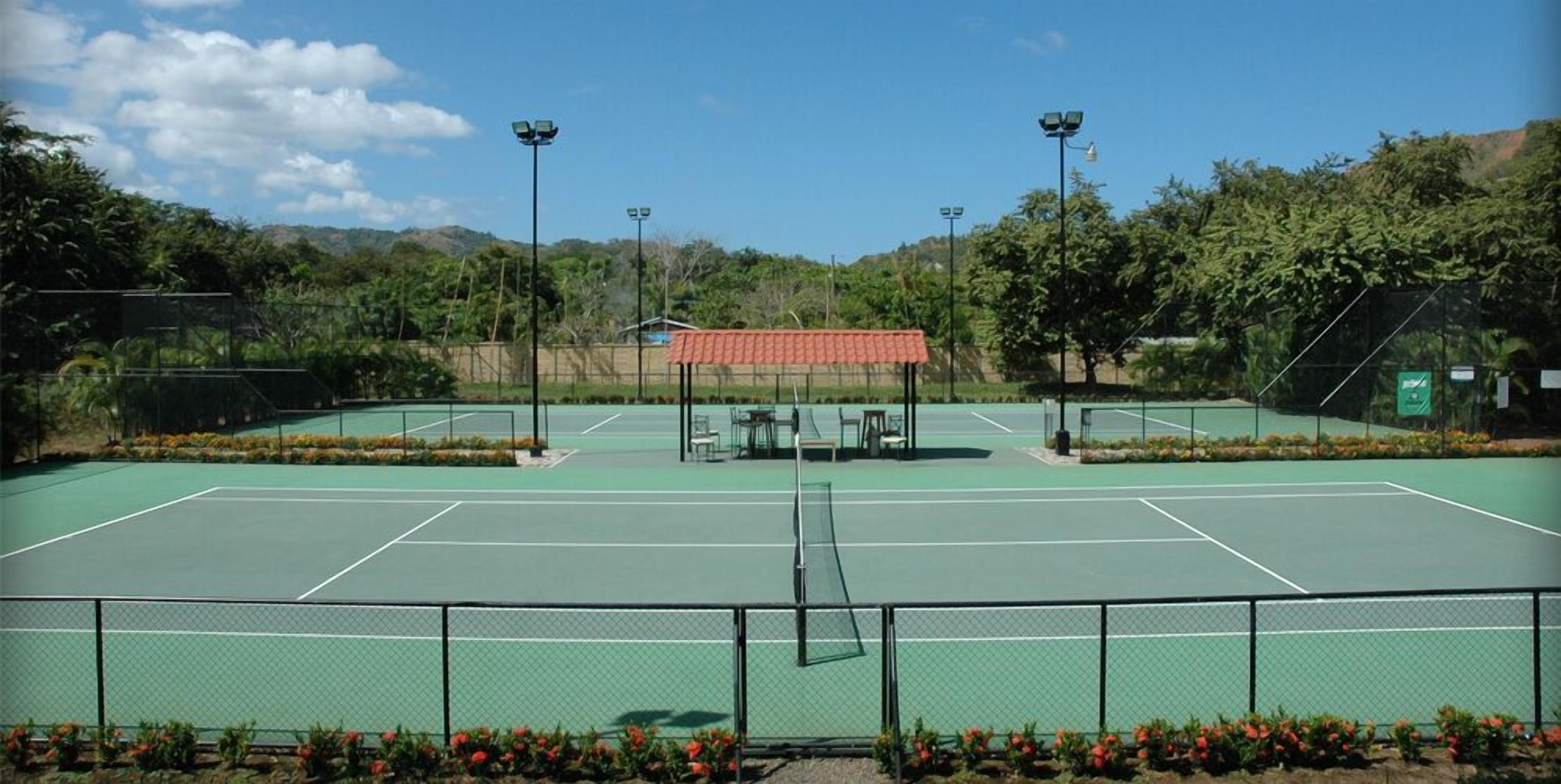 A tennis court with four light poles, surrounded by greenery and a small building with a red roof.