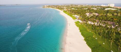 An aerial view of a coastal area with a beach, a boat trail, and a residential area with houses and palm trees.
