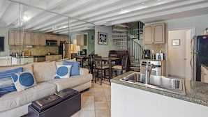 A modern kitchen with a built-in island, a dining area with a spiral staircase, and a living room with a sofa and ottoman.