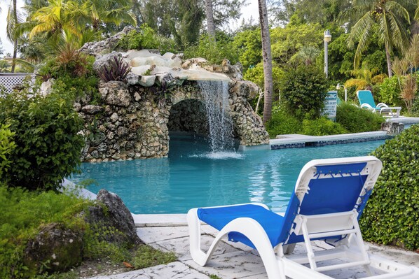 Piscina con cascada, rodeada de exuberante follaje y una silla de descanso.