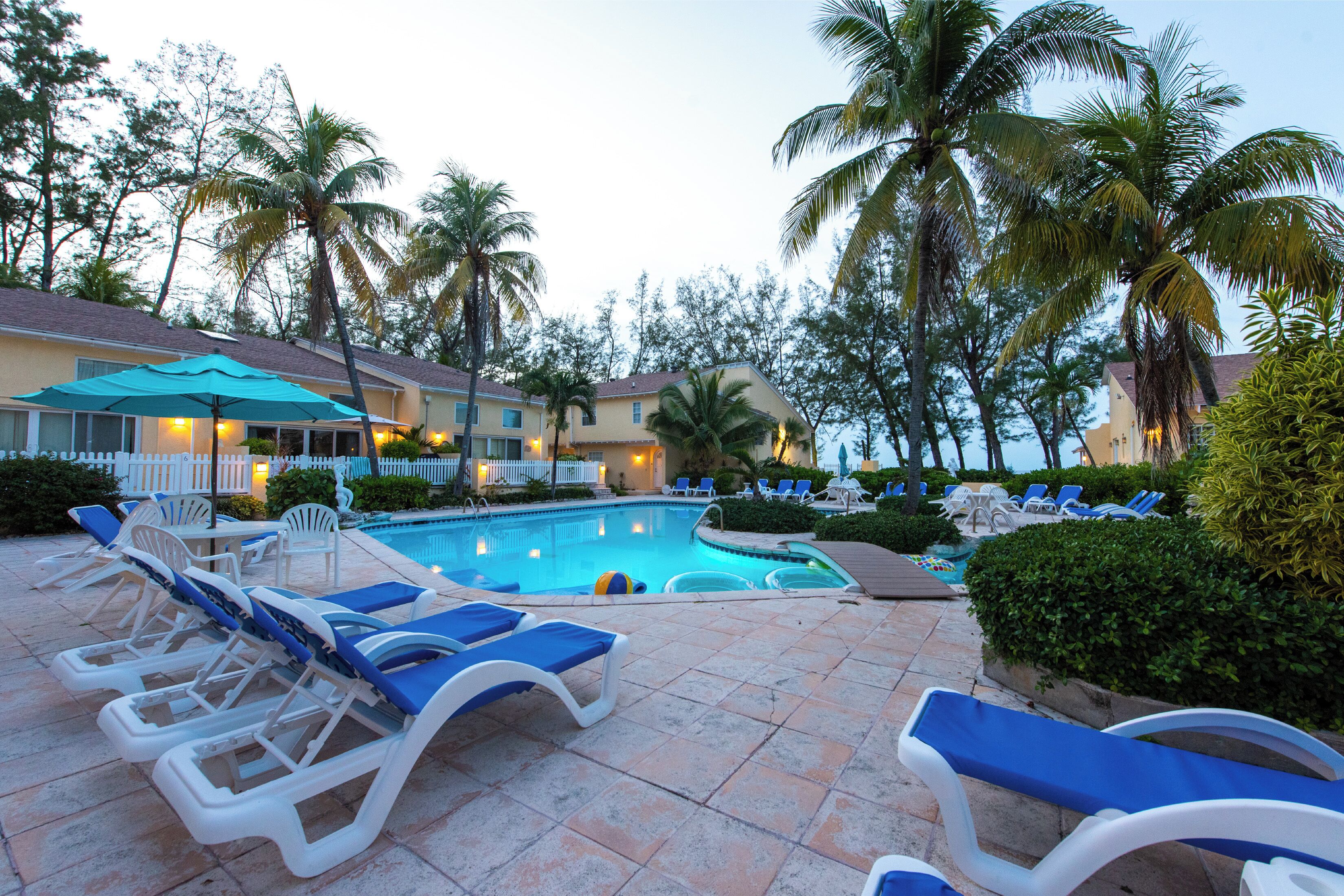 A pool area with lounge chairs and umbrellas, surrounded by palm trees and a building with a tiled roof.