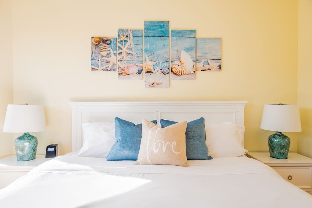 Una cama bien tendida con ropa de cama blanca y almohadas azules y beige, un cuadro de conchas marinas y estrellas de mar, y dos lámparas verdes sobre mesitas de noche.