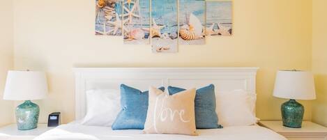 Una cama bien tendida con ropa de cama blanca y almohadas azules y beige, un cuadro de conchas marinas y estrellas de mar, y dos lámparas verdes sobre mesitas de noche.