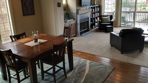 Een ruime woonkamer met een houten eettafel en stoelen, een open haard, een boekenkast en een comfortabele zithoek met een bank en fauteuils.