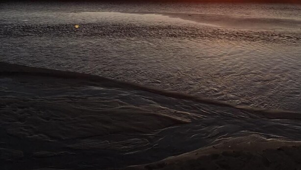 Een zonsondergang aan het strand, met een rustige zee en verafgelegen rotsformaties.
