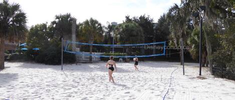 A sandy beach volleyball court with a net, surrounded by palm trees and umbrellas.