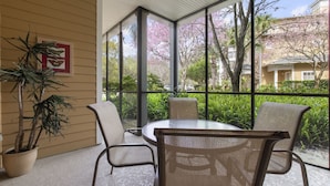 Un patio techado con una mesa redonda y cuatro sillas, ventanas amplias que ofrecen vistas a árboles y un edificio.
