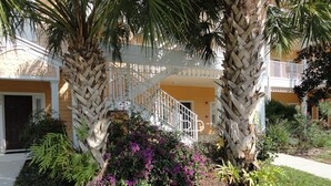 Um edifício de dois andares com guarda-corpo de varanda branco, cercado por vegetação exuberante e plantas floridas.