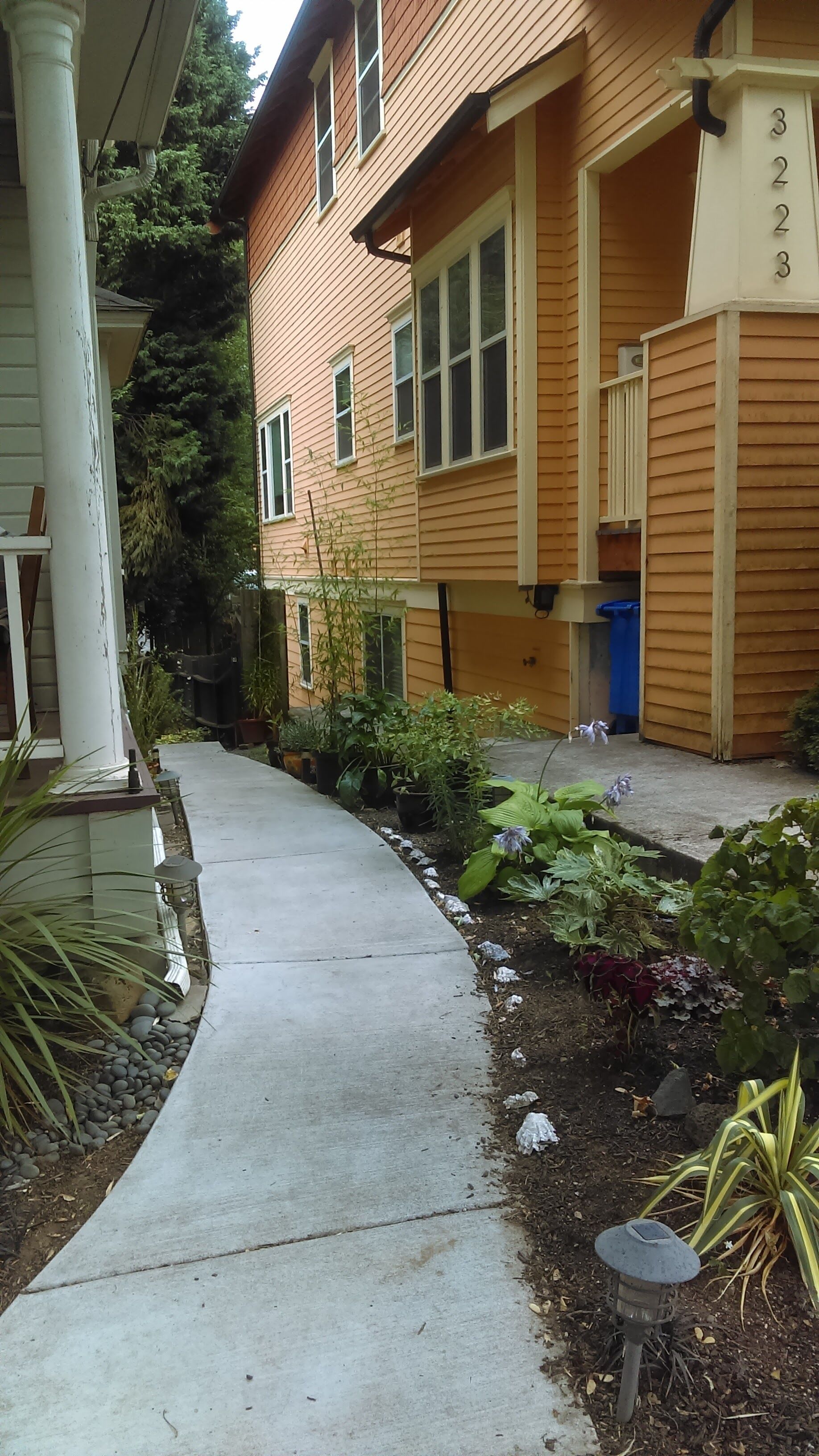 A residential building with a covered porch, a concrete pathway, and a garden area with various plants and a small decorative lantern.