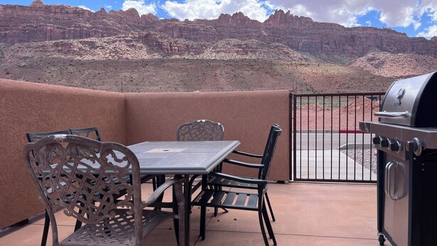 An outdoor patio with a table and chairs, a grill, and a view of a mountainous landscape.