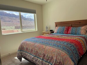 A bedroom with a large window, a wooden headboard, a colorful bedspread, and a nightstand with a vase and a lamp.