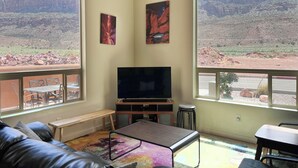 A living room with a large TV, a couch, a coffee table, and a view of a desert landscape through large windows.