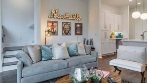 En moderne stue med en grå sofa, et sofabord med dekorative genstande, og et køkkenområde med hvide skabe og en hængende lampe.
