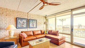 A modern living room with a brown leather sectional sofa, a glass coffee table, and a ceiling fan. There are two framed artworks on the brick wall, a wicker side table with a lamp, and a view of the outside balcony.