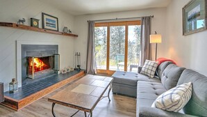 A cozy living room with a fireplace, a sofa, a coffee table, and a large window with a view of trees. - Remodeled Condo Near Slopes, Pool/Hot Tub Access! (Silverthorne)