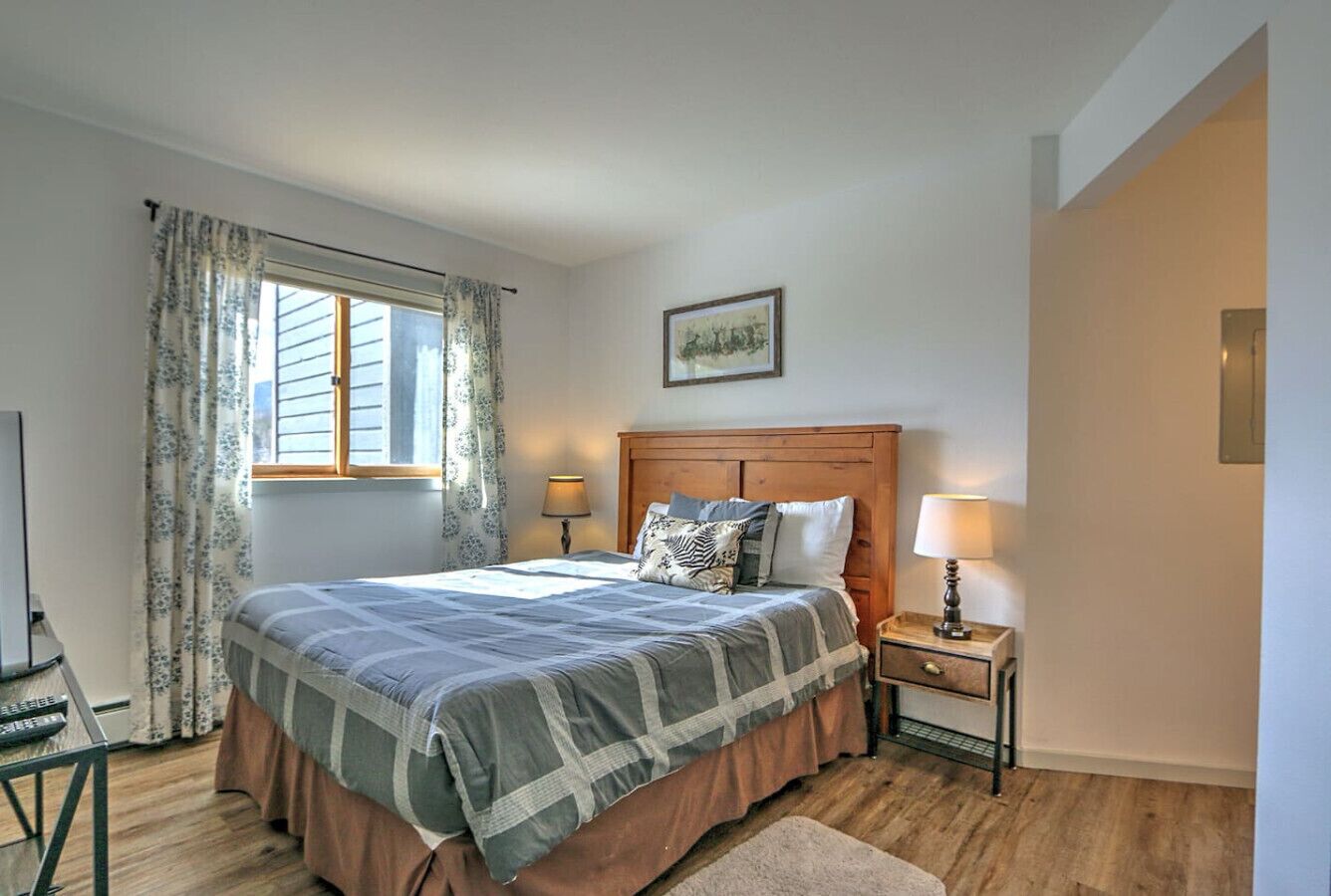 Ein Schlafzimmer mit einem Bett, zwei Nachttischen, einem fenster mit Vorhängen, einem gerahmten Bild an der Wand und einem Holzboden.