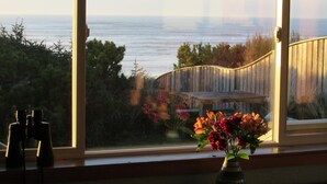 Vista a través de una ventana con una terraza de madera, un florero y unos binoculares en el alféizar.