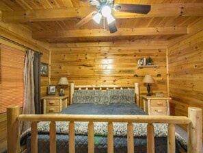 Een slaapkamer in een houten blokhut met een bed, twee nachtkastjes, een plafondventilator en een raam met jaloezieën.