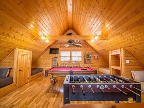 Une pièce mansardée chaleureuse avec des murs en bois et un plafond en pente. Elle est équipée d’une table de billard, d’une table de baby-foot et d’une fenêtre qui laisse entrer la lumière naturelle.