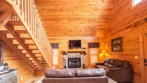 L’intérieur d’une cabane en rondins, avec une cheminée en pierre, un canapé, un téléviseur et un escalier.