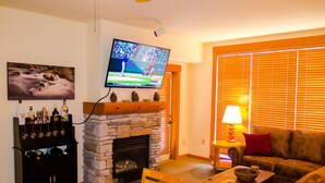 A living room with a stone fireplace, a television mounted on the wall, a wooden dining table, and a sofa.