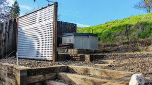 Eine Holzterrasse mit einem Gitterzaun, eine kleine Gartenhütte und ein grasbewachsener Hügel im Hintergrund.