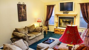 Ein Wohnzimmer mit Sofa, Sessel und Hocker, einem Fernseher über einem Kamin und einer Lampe auf einem Beistelltisch.