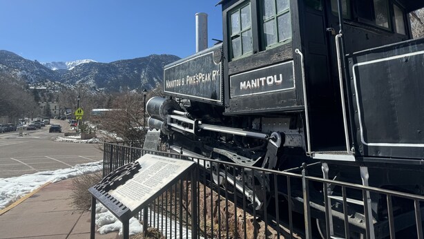 Una locomotora negra con letreros que indican 'Manitou & Pike's Peak' y 'Manitou', frente a un paisaje montañoso cubierto de nieve.