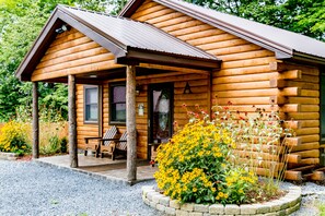 A log cabin with a porch, a bench, and a flower bed. - Log Cabin Retreat, Cayuga Wine Trail, Dogs Welcome, Open Year-round (Ovid)
