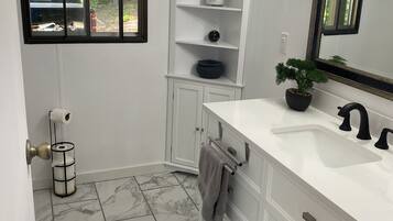 Une salle de bain avec un meuble-lavabo blanc, un grand miroir, une plante en pot et une fenêtre donnant sur l’extérieur.