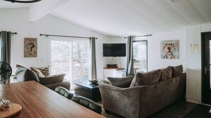 Amplia sala de estar con una mesa de comedor de madera, asientos cómodos, un televisor de pantalla plana y ventanas grandes que ofrecen vistas al exterior.