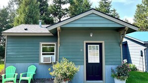 A small, single-story house with a dark blue front door, a small porch with two green Adirondack chairs, and two potted plants. - Cozy Cabin With Mountain Views (Columbia Falls)